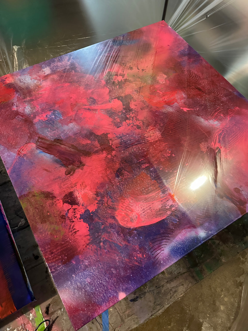 An abstract square canvas painting with vibrant red, pink, purple, and blue brushstrokes, photographed from an angle in what appears to be an art studio or workshop.